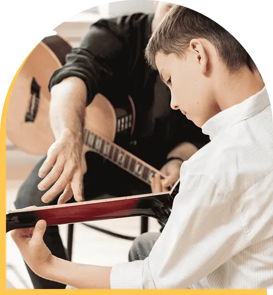 A boy in a white shirt learns to play guitar during music lessons in Westchase, guided by an adult nearby who points at the strings and offers encouragement.