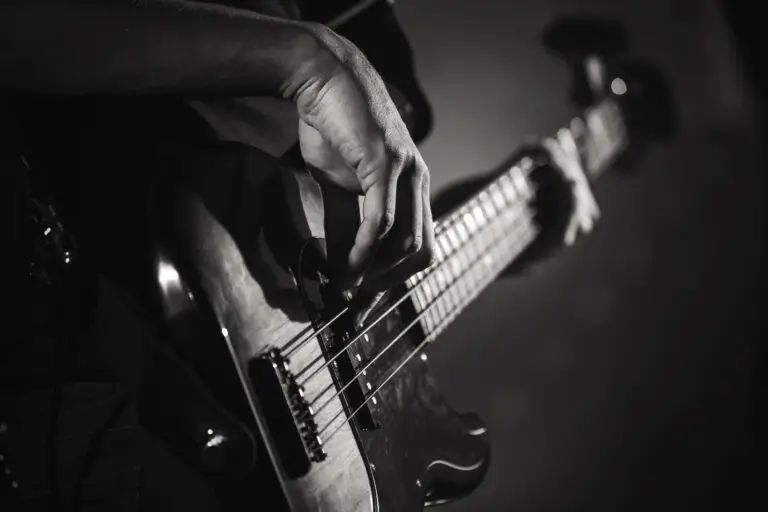 A close-up, black and white photo of a person’s hand playing an electric bass guitar, focusing on the fingers plucking the strings and part of the instrument's neck and body—a striking image that captures the artistry taught at Lehi School of Music.
