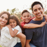 Young Hispanic parents piggyback their children in the park, smiling to camera, focus on foreground